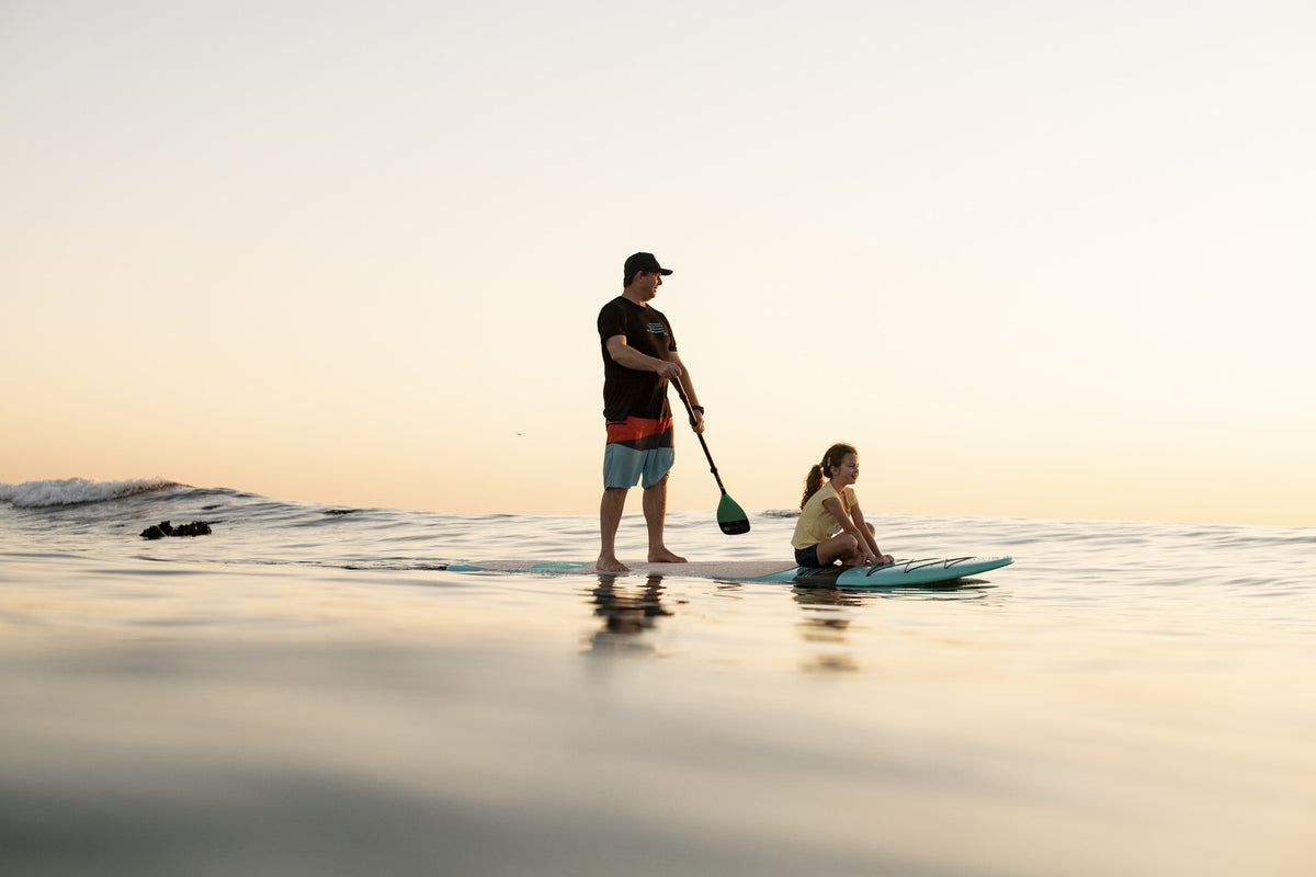 A paddler standing on an RYD stand up paddleboard at sunset, with a young girl sitting on the front of the board