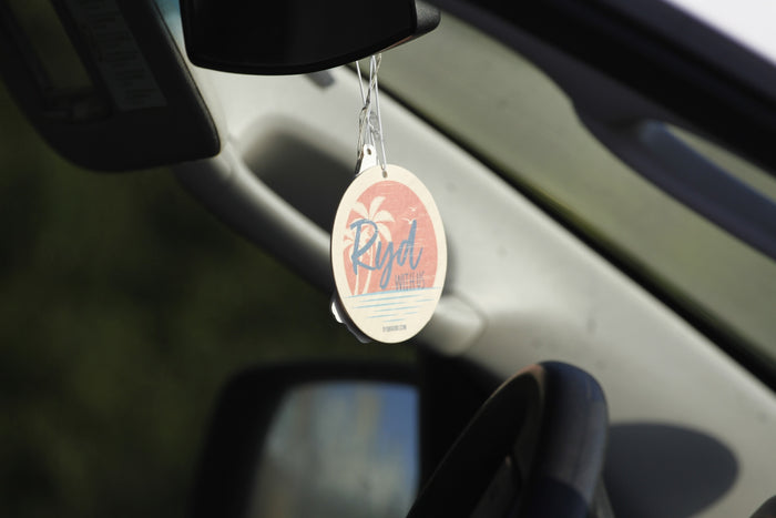 RYD coral air freshener with palm tree silhouette and 'Ryd With Us' branding, hanging from a rearview mirror
