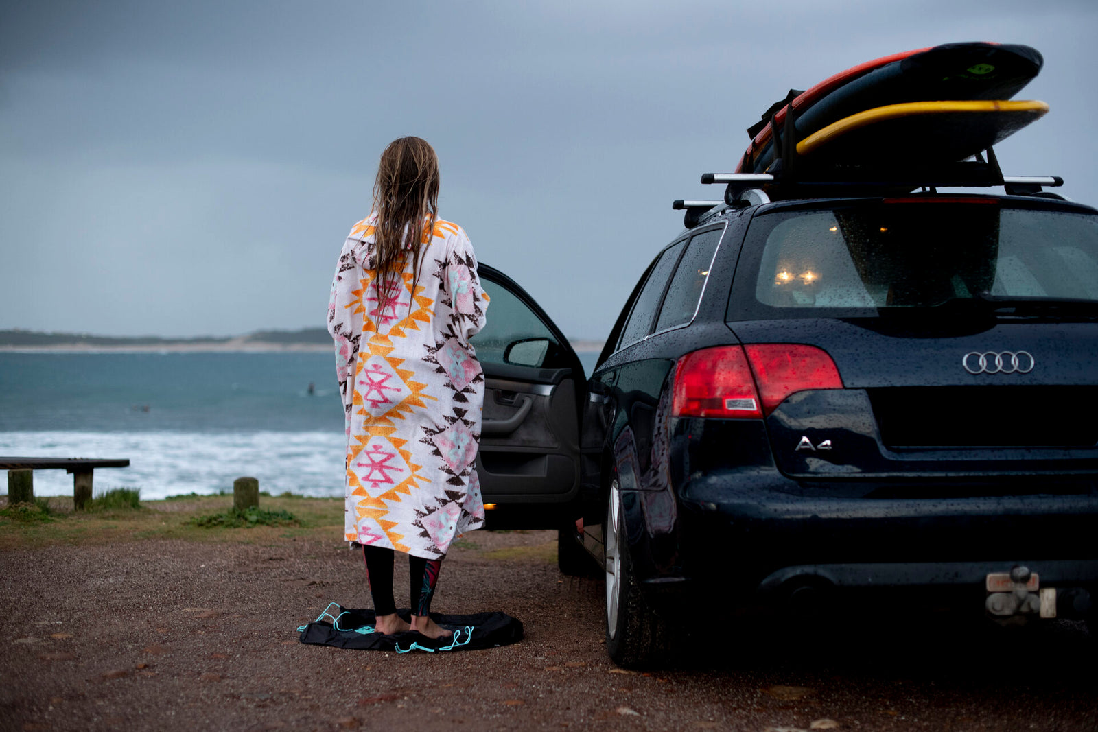 A woman wrapped in a colourful changing towel standing beside a car loaded with surfboards, looking out at the ocean under a stormy sky