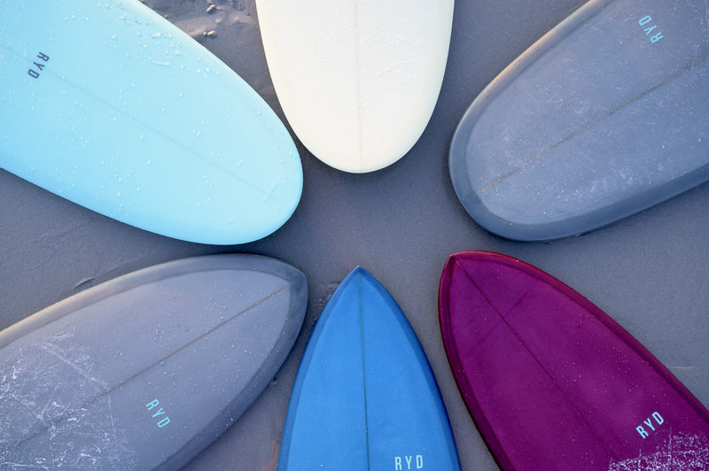 Six RYD surfboards in different jewel-tone colourways arranged in a star pattern on wet sand, each stamped with the RYD logo