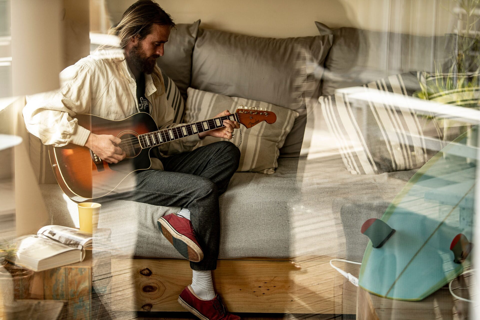 A bearded man sitting on a couch at home playing an acoustic guitar, with a surfboard visible in the reflection in the foreground