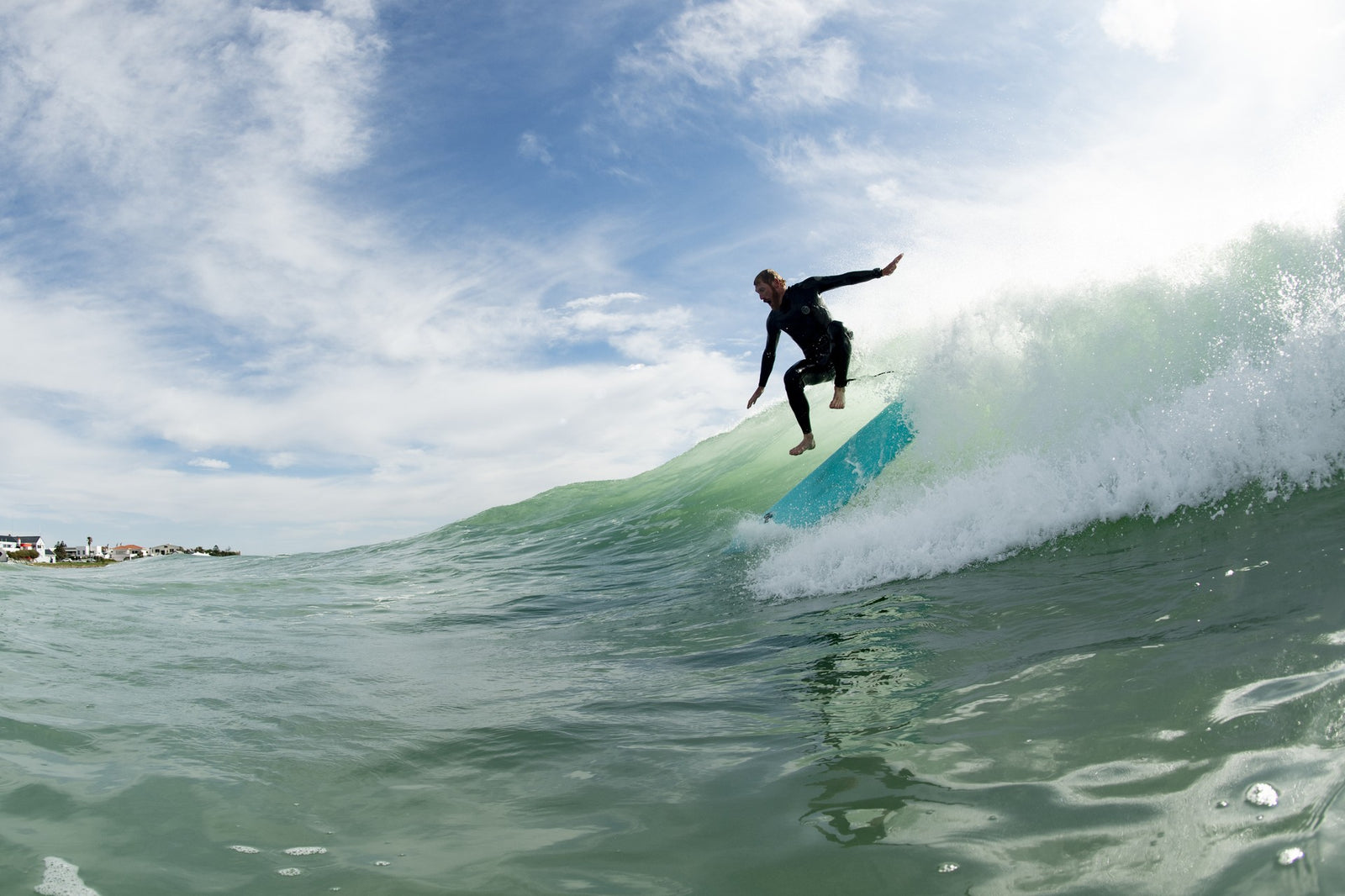 A surfer catching air above a breaking wave on a teal surfboard, silhouetted against a bright sky with coastal houses visible