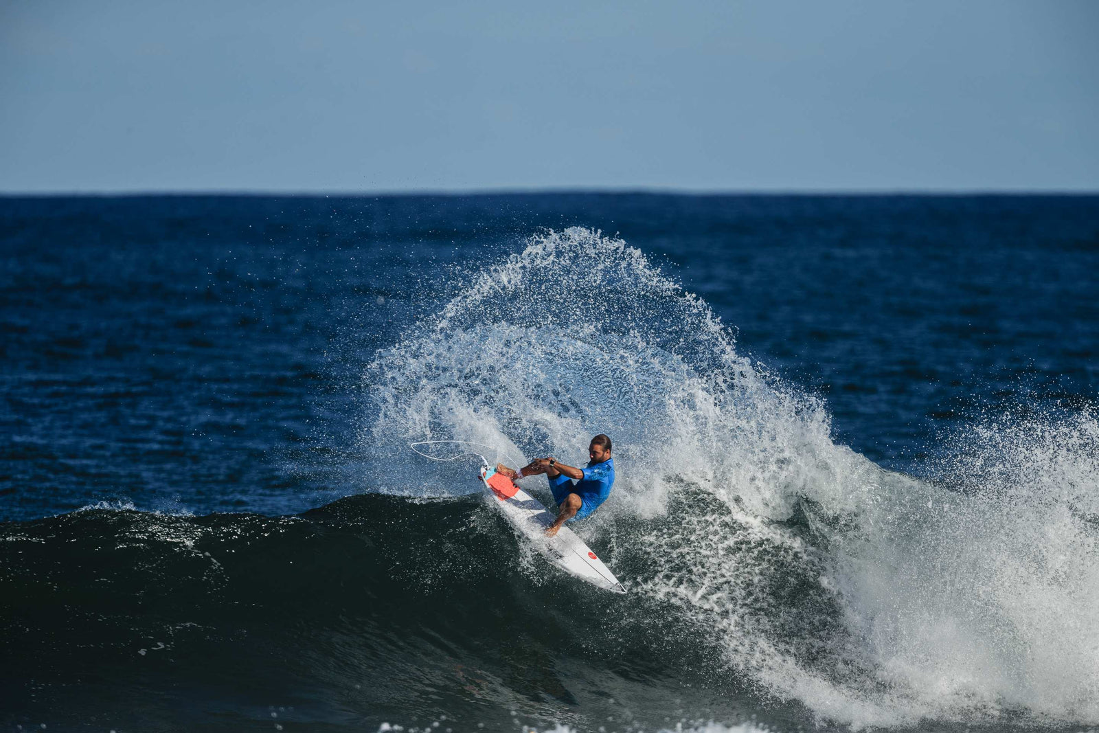 A surfer in a blue rash vest carving hard off the top of a wave on a surfboard with an orange traction pad